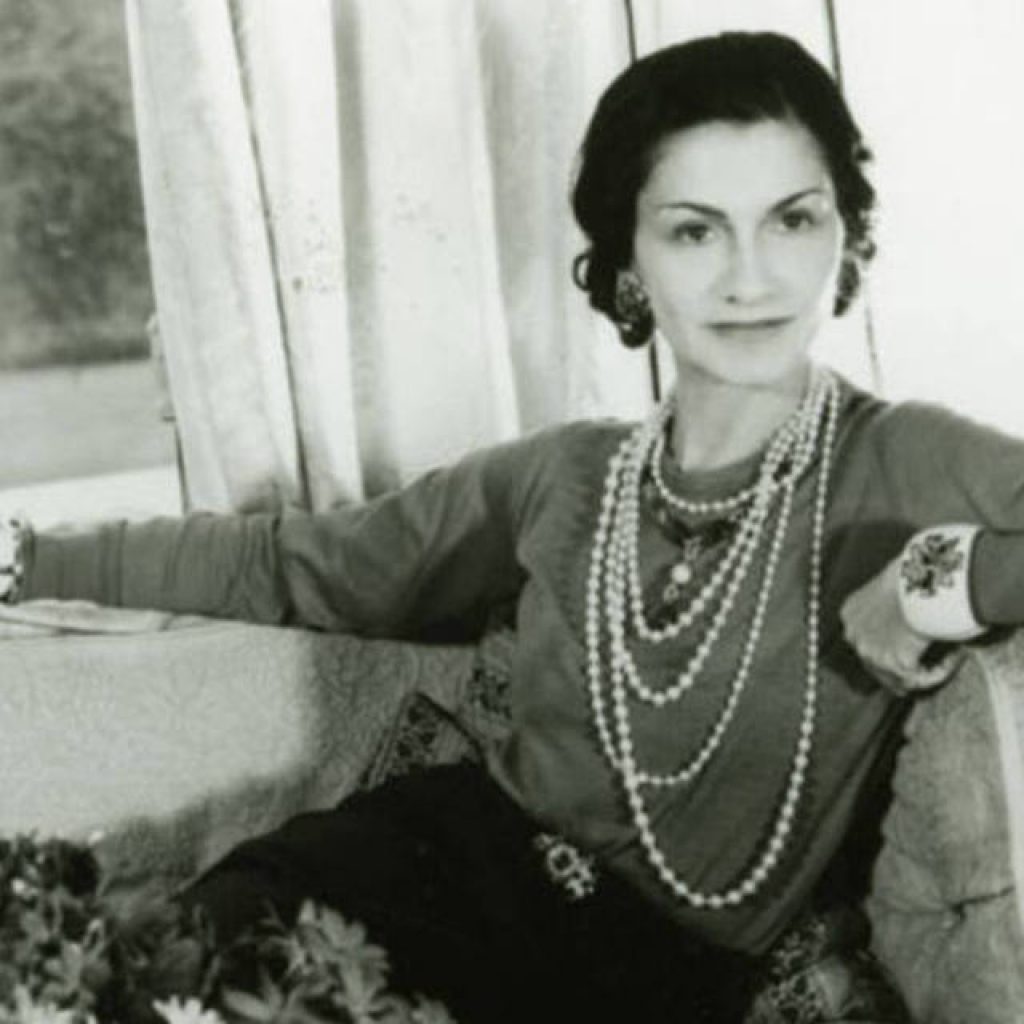 Foto em preto e branco de Coco Chanel em um sofá com blusa de jersey, colar de perolas e pulseiras iconicas